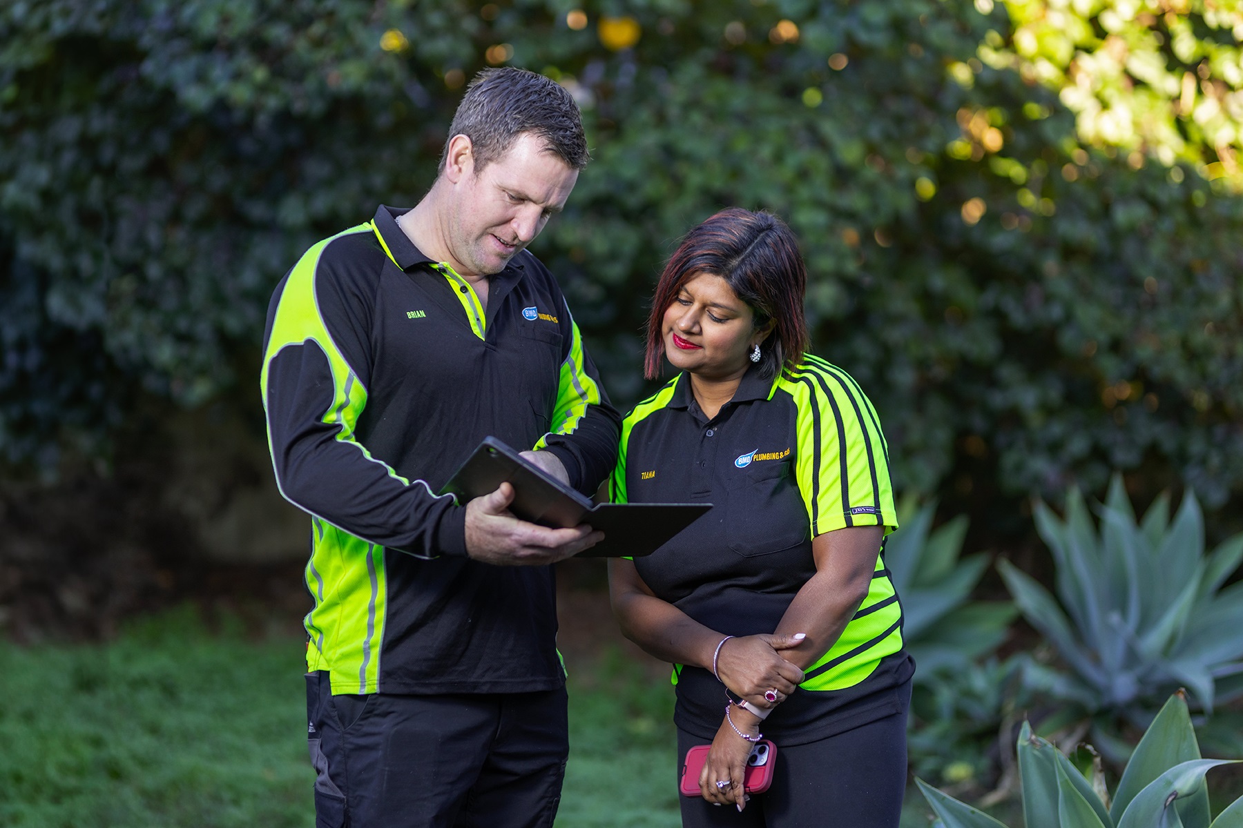 Photo of Brian and Tiana McDaid, owners of BMD Plumbing and Gas. They are standing in a garden, looking at a tablet device.  