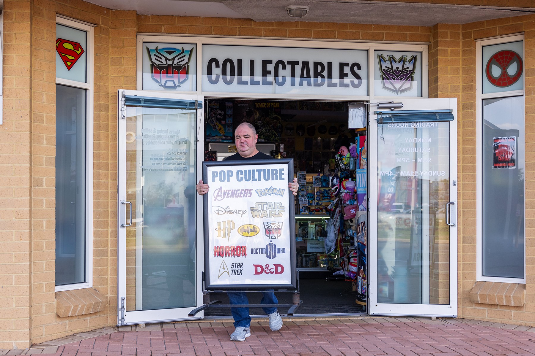 Photo of Chris Topher, owner of Collector Zone, in front of his business premises in Clarkson. He is carrying a sign out to the front of the store.