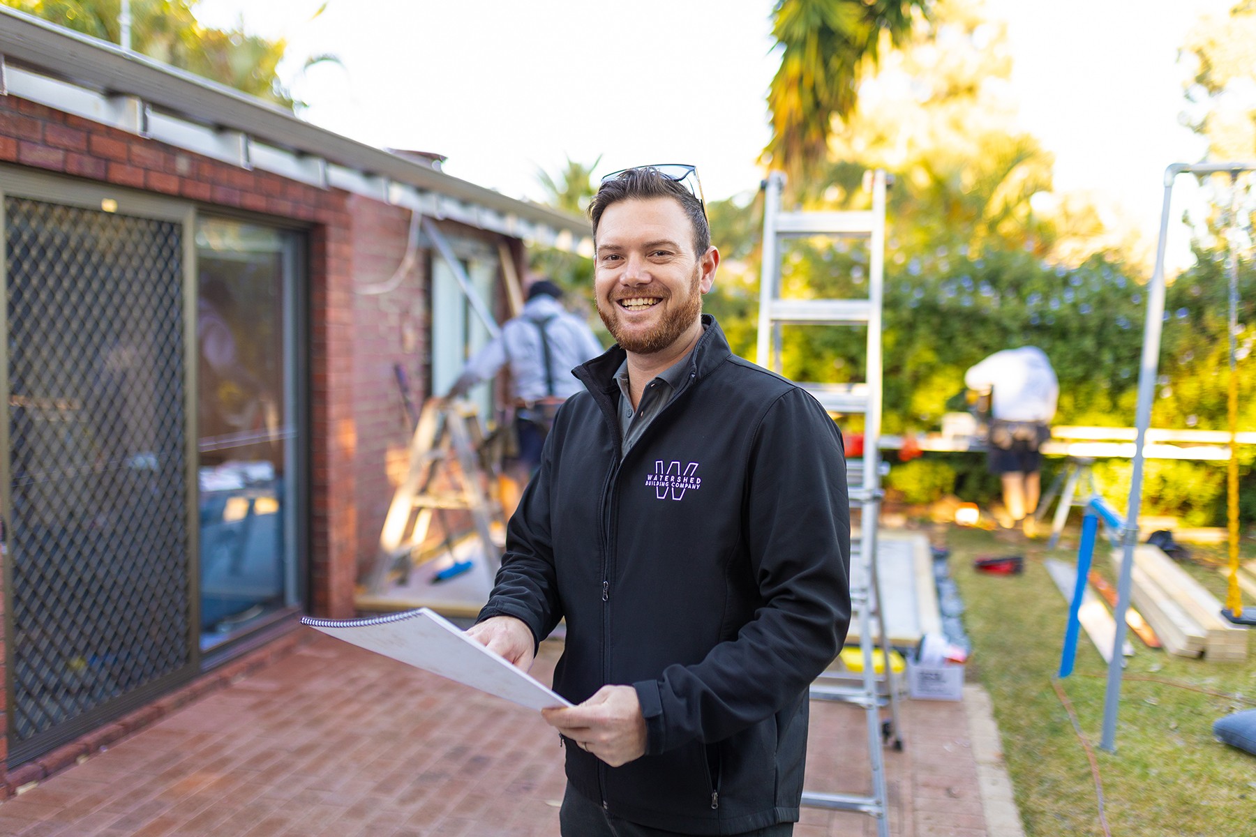 Evan Turners, owner of Watershed Building Company, posing on a building site whilst people work in the background.
