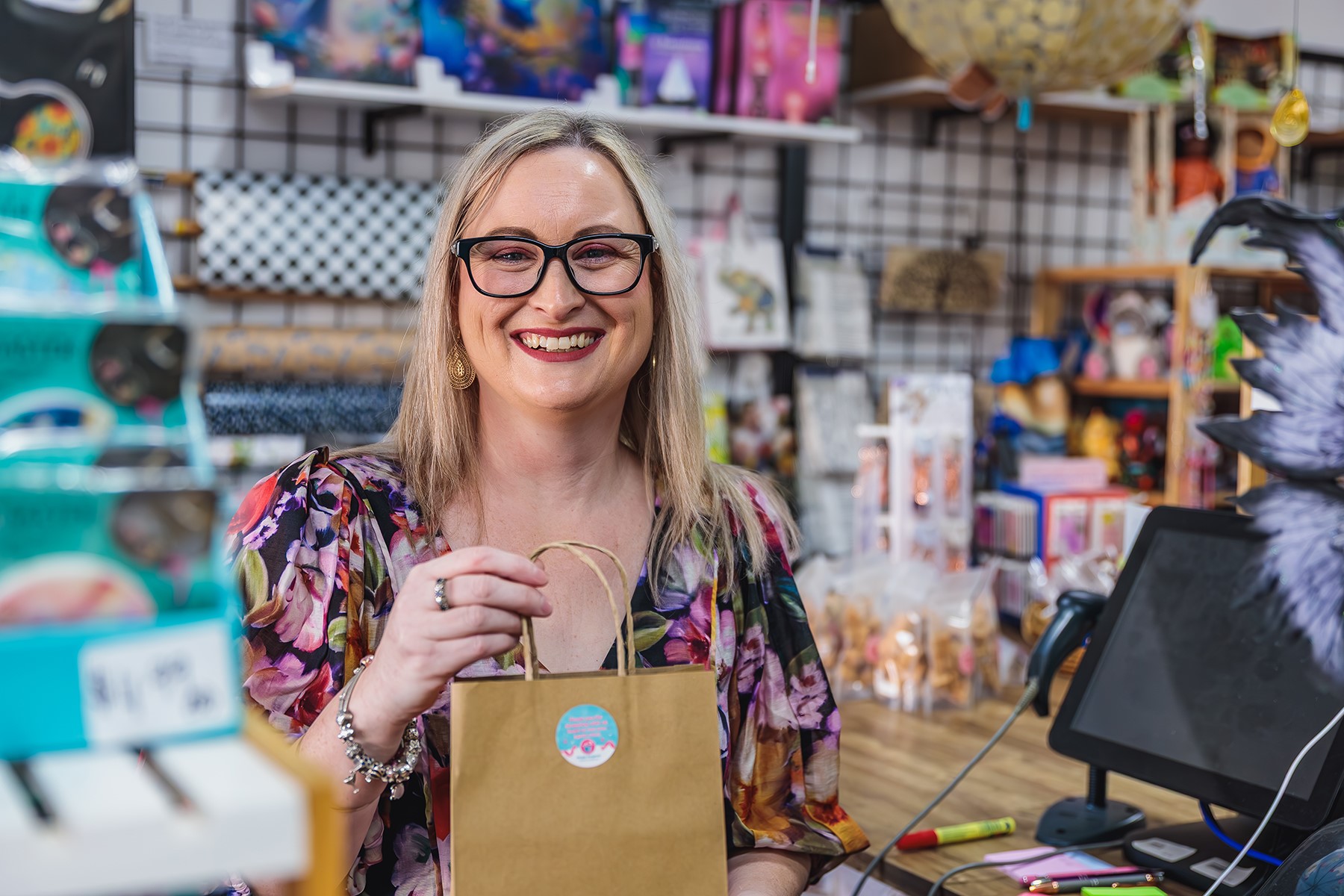 A woman standing at the counter of a retail store. She is holding a bag and smiling at the camera.