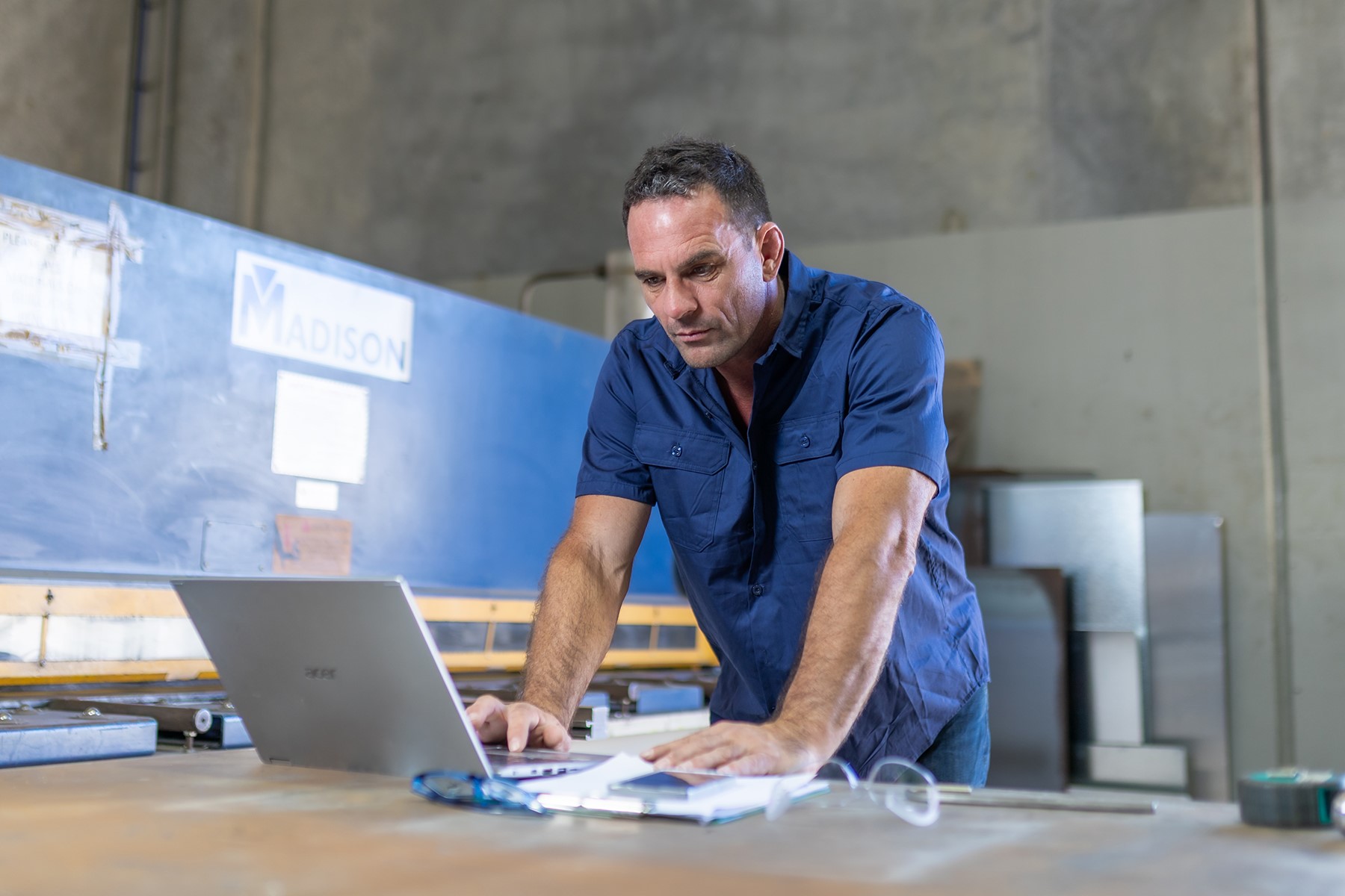 A man standing in an industrial workshop. He is standing at a workbench, reading from a laptop screen.