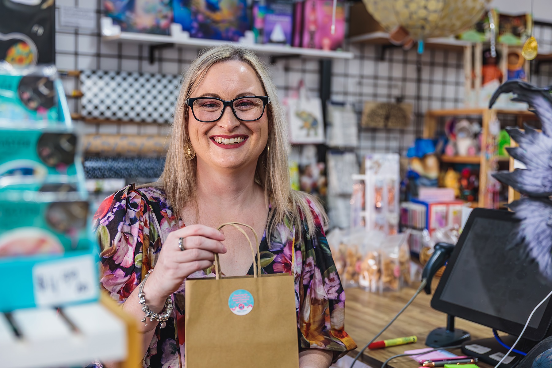 Photo of a female business owner in her retail store. She is standing behind the counter, holding a small paper gift bag.