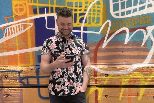 Photo of a man looking at his smart phone. He is standing in front of a wall covered with street art.