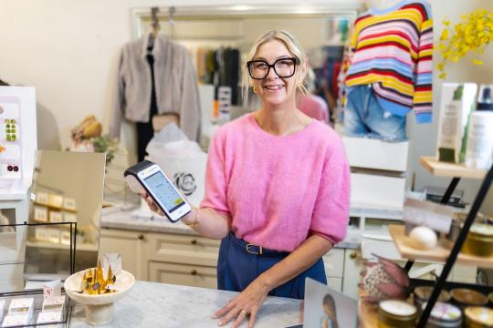 Photo of a female business owner. She is standing in her retail clothing business and is holding an EFTPOS terminal.