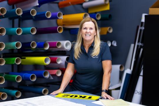 Photo of a woman standing in a signage shop. She is stranding in front of a rack with rolls of vinyl on display.