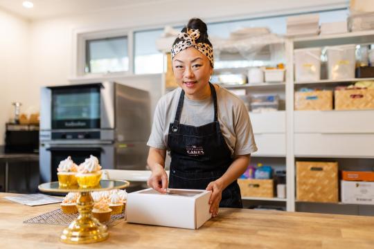 A woman standing at the counter of her cake business. There are cupcakes on display and she is holding a box with cakes.