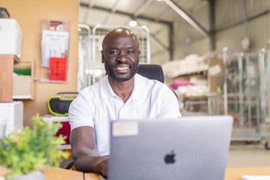 Photo of a man sitting at a desk in his laundry business. He is working on a laptop.