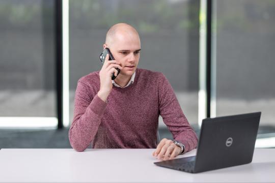 Photo of a man sitting at a desk. He is speaking on the telephone and looking at a laptop screen.