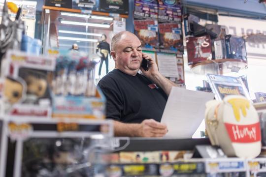 Photo of a male business owner in his comic book store. He is standing behind the front counter, reading from a piece of paper while talking on the phone.