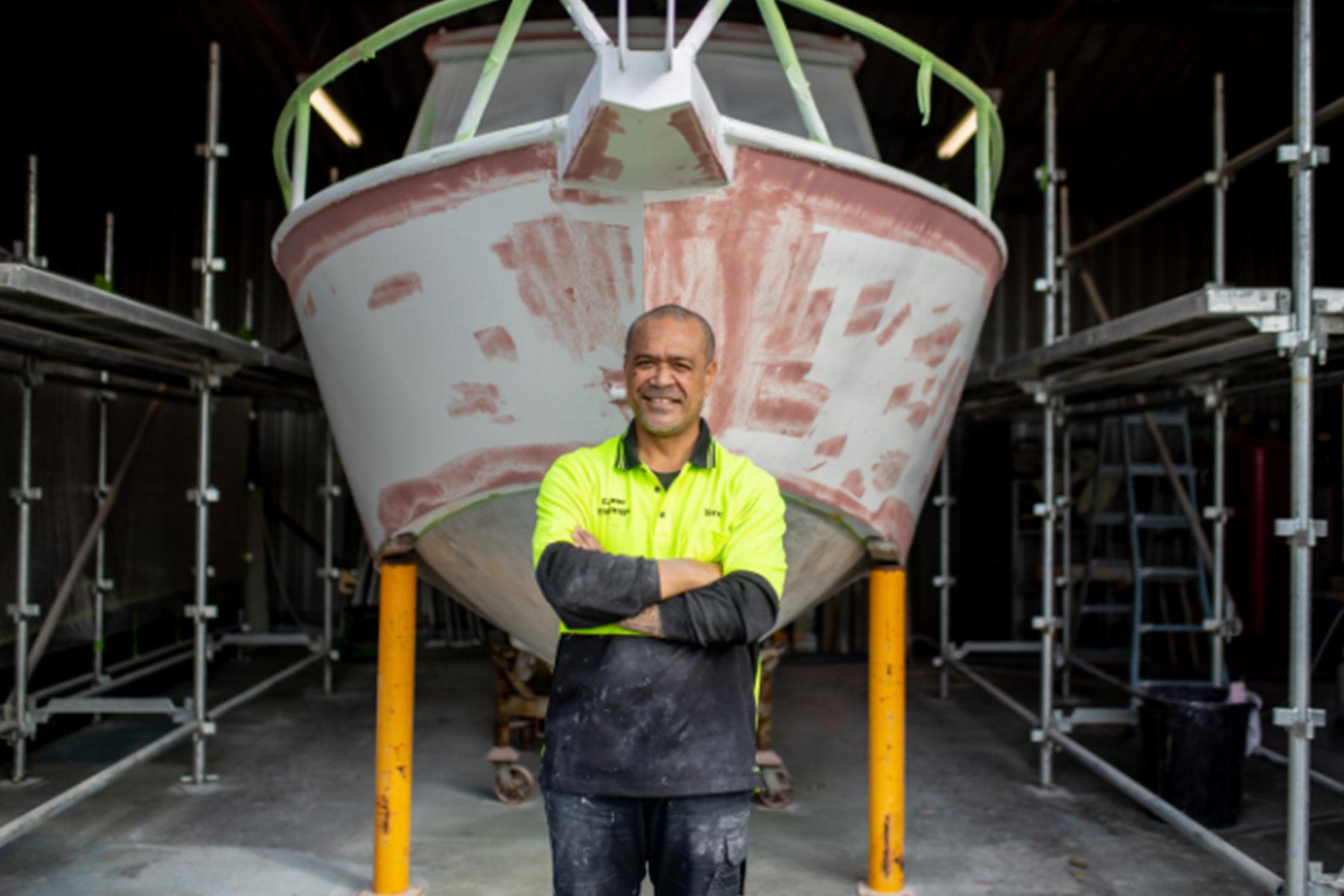 Image shows a man standing in front of a boat with his arms crossed smiling at the camera