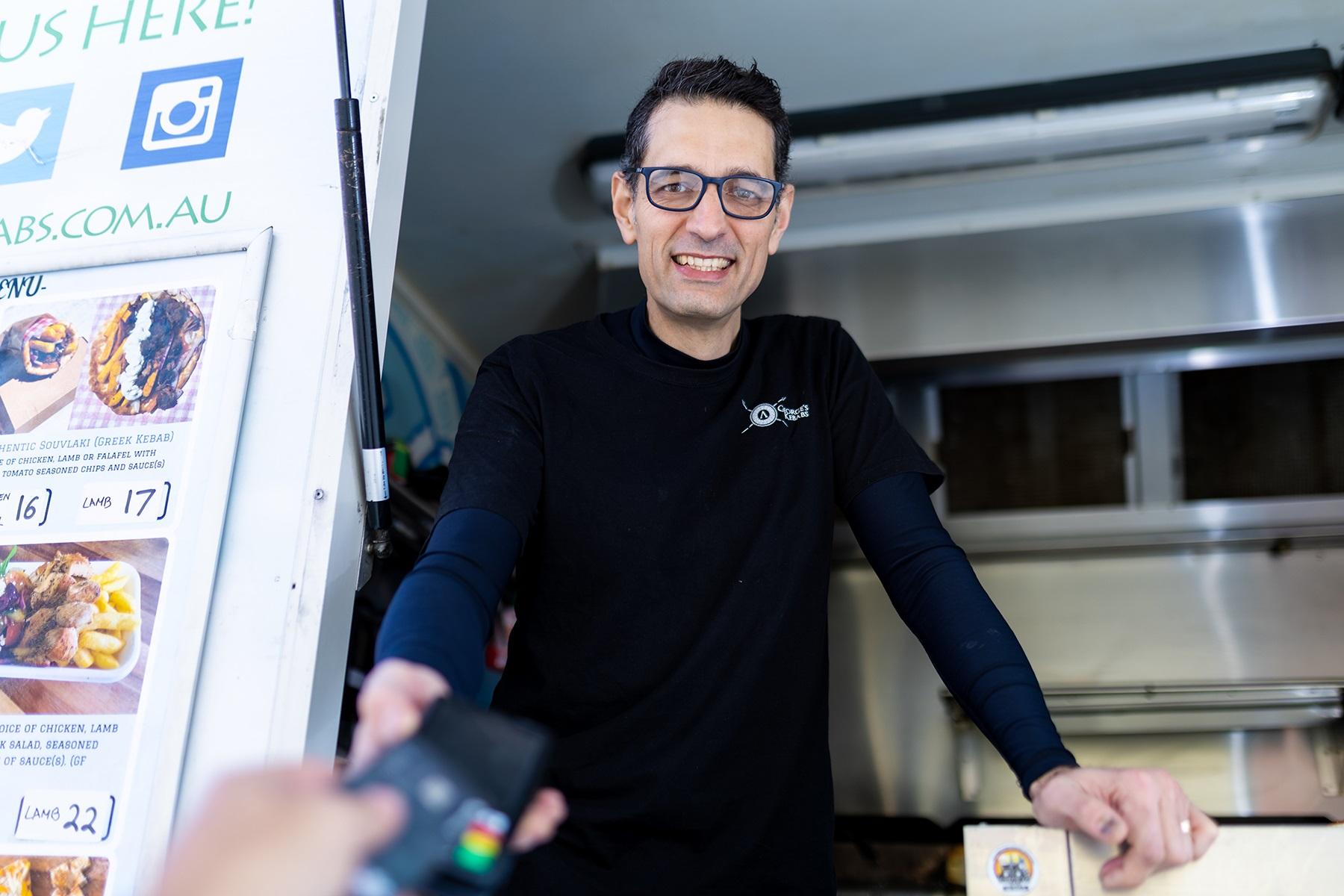 Photo of a food truck owner. He is standing in his food truck while a customer pays using a credit card.