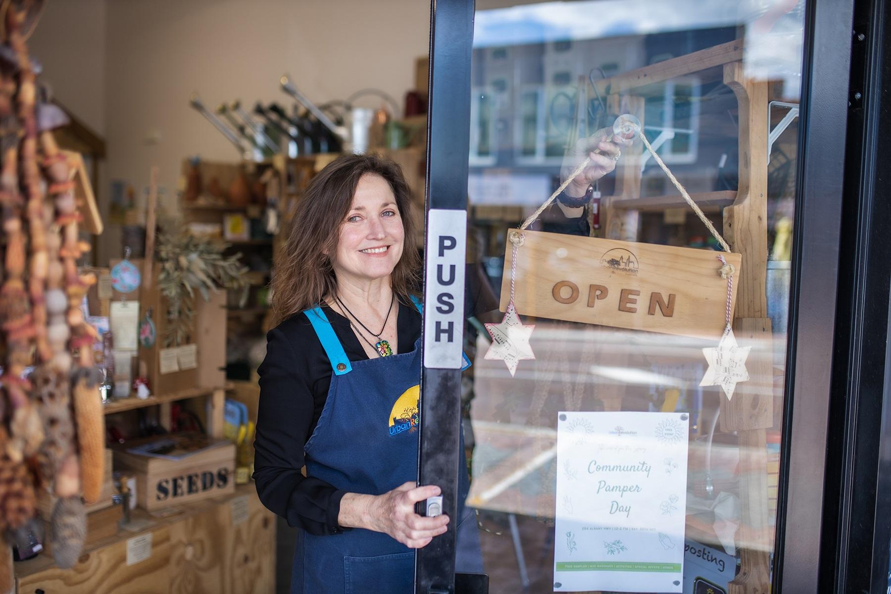 Photo of a female business owner. She is at the door of her retail store and is hanging up an open for business sign.
