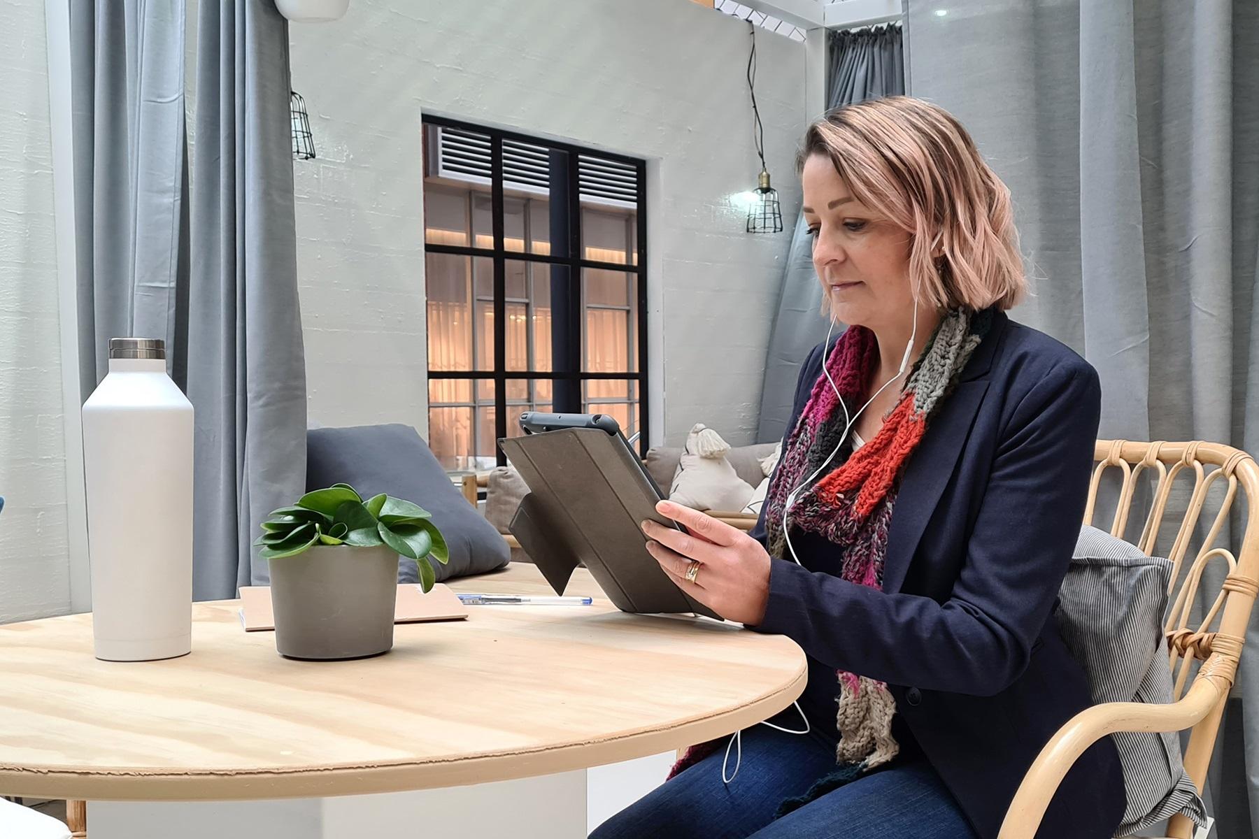 Photo of a woman sitting at a dining table. She is reading from a tablet device.