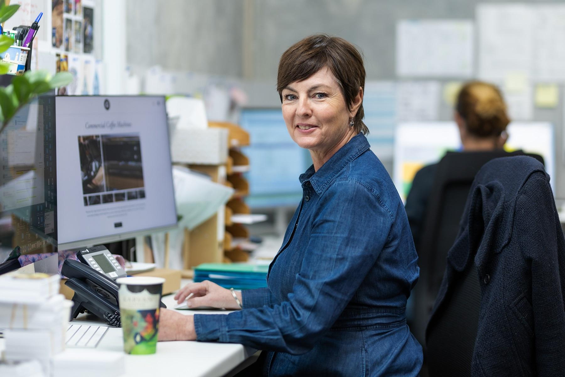 Photo of a female business owner. She is sitting at a desk, in front of a computer, and smiling at the camera.