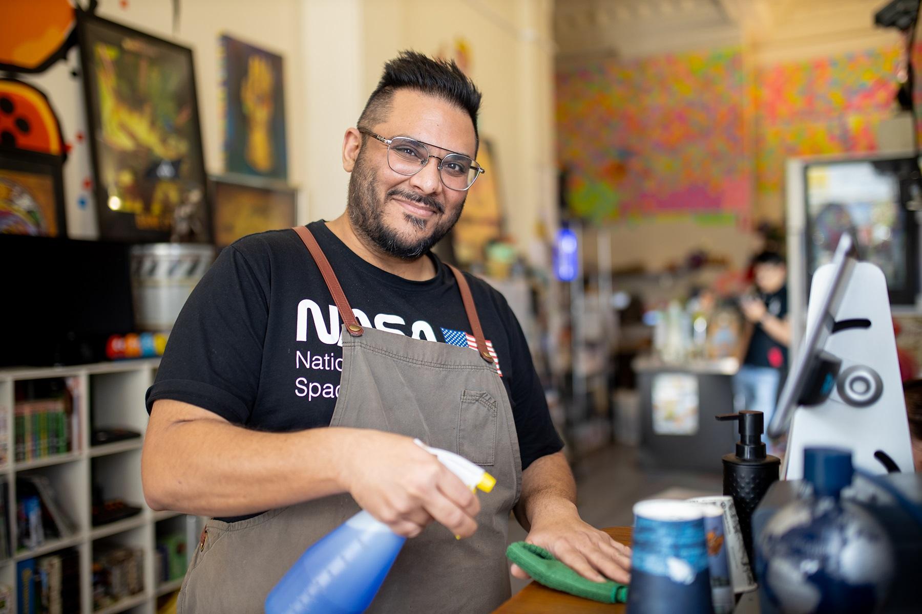 Photo of a male cafe owner. He is wiping down the area in-front of a coffee machine.