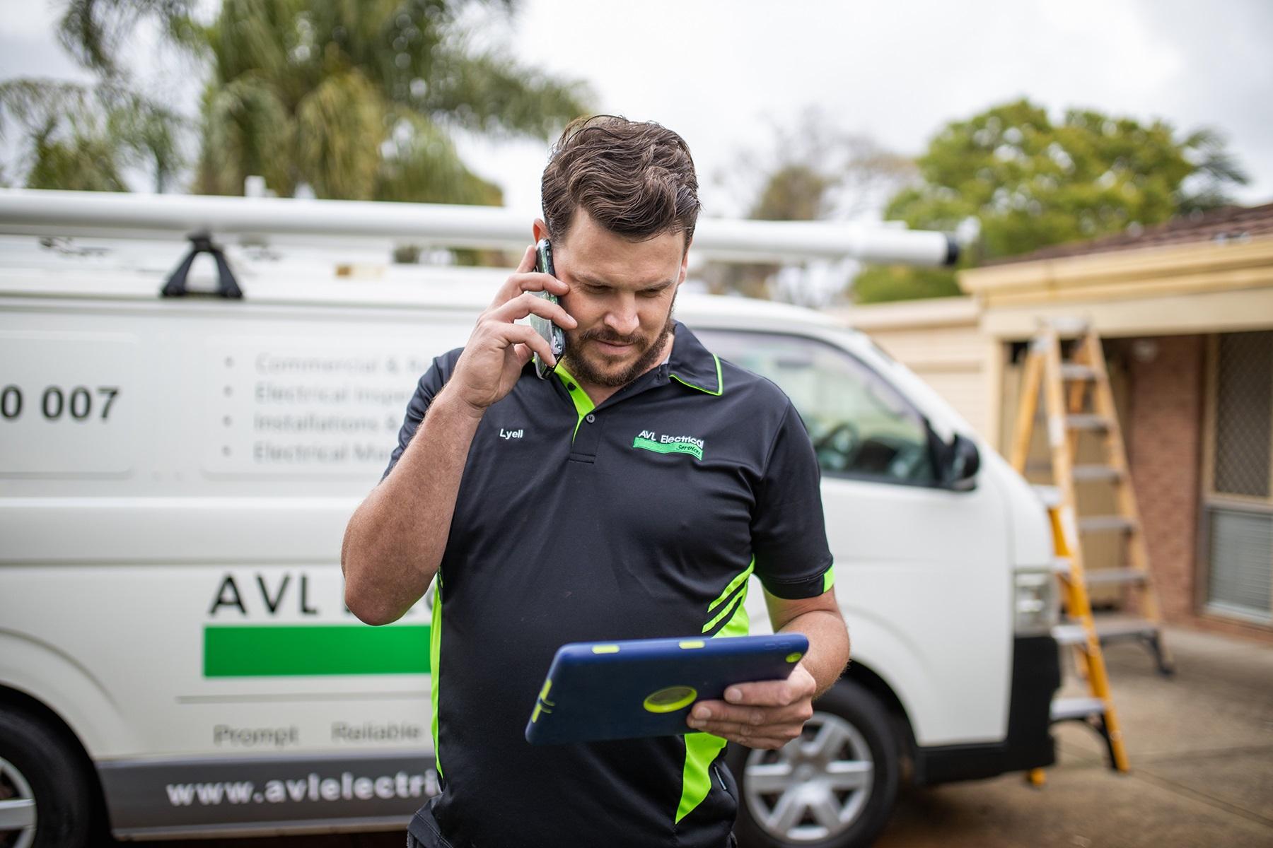 Photo of a male tradie. He is standing in front of his work van while he speaks on his smartphone and reads from a tablet device.