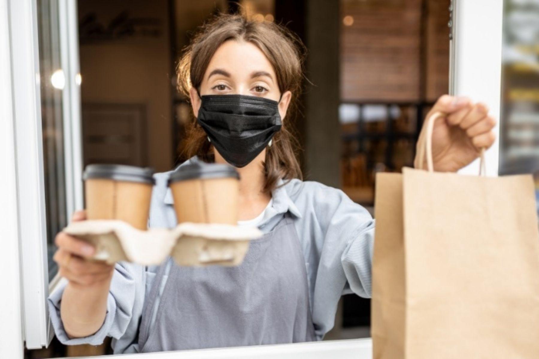 A photo of a masked cafe worker handing coffee and food to a customer through a take away window.
