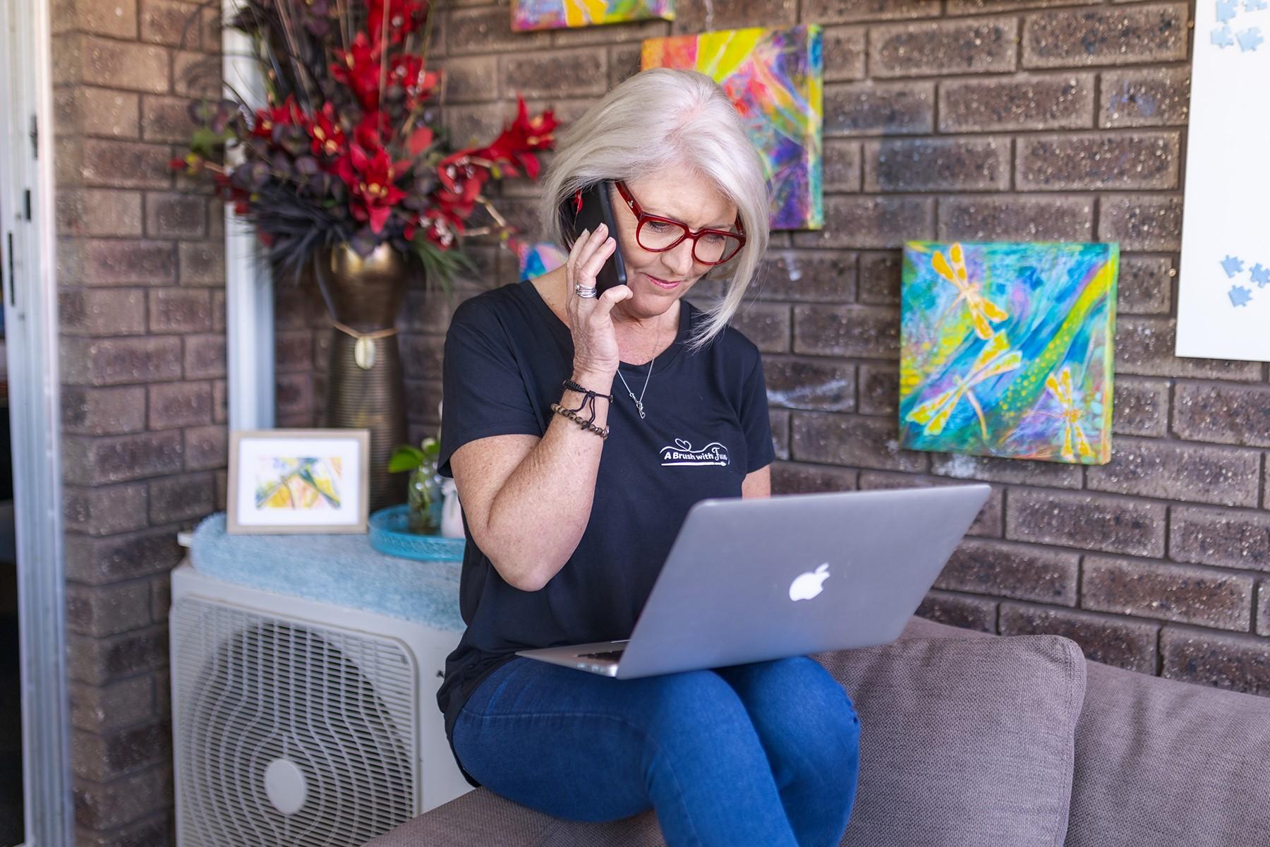 A woman sitting on the arm of a couch. She is looking at a laptop while speaking on a smart phone.