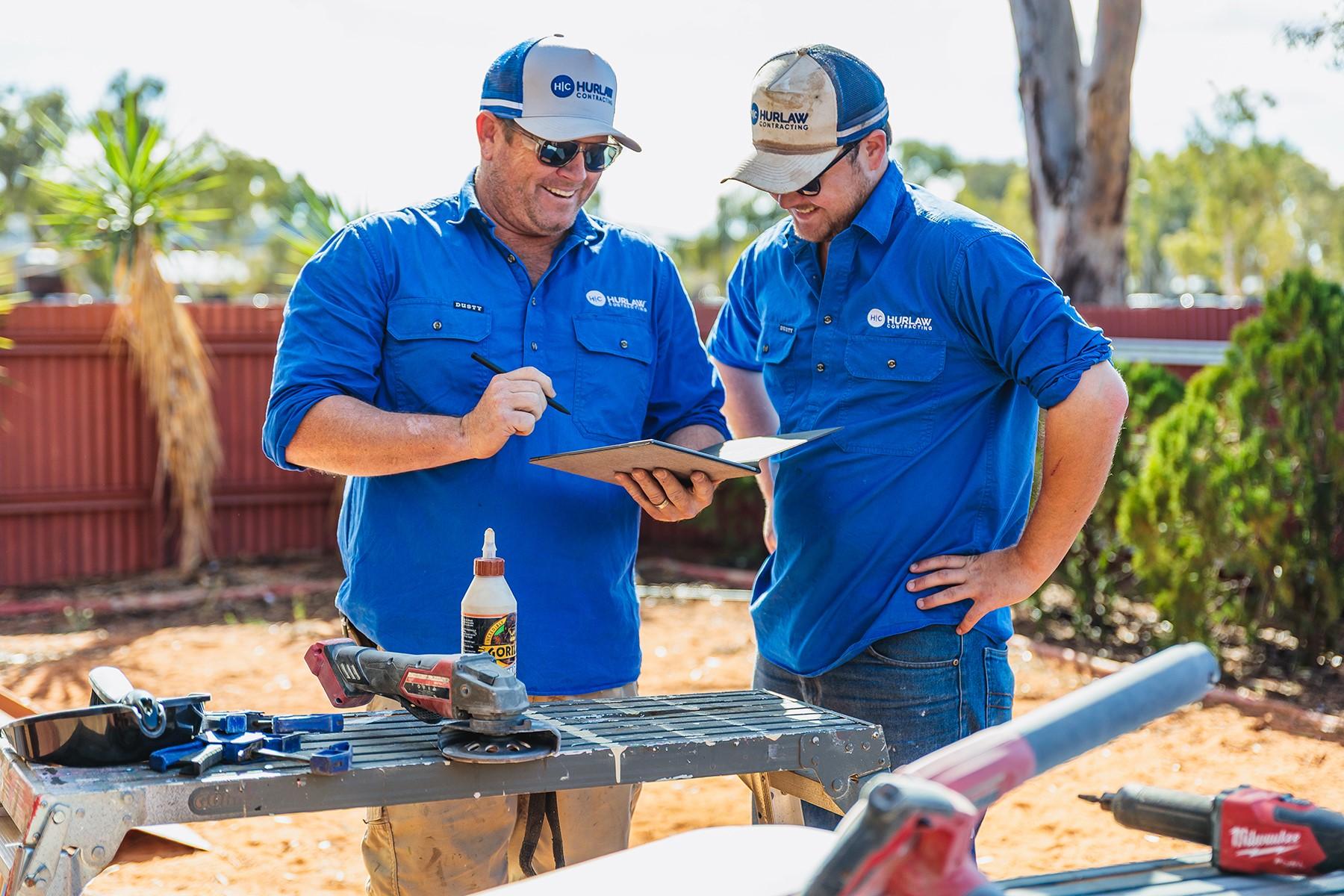 Two tradesmen reviewing a document. They are standing in a residential property with a workbench and tools.