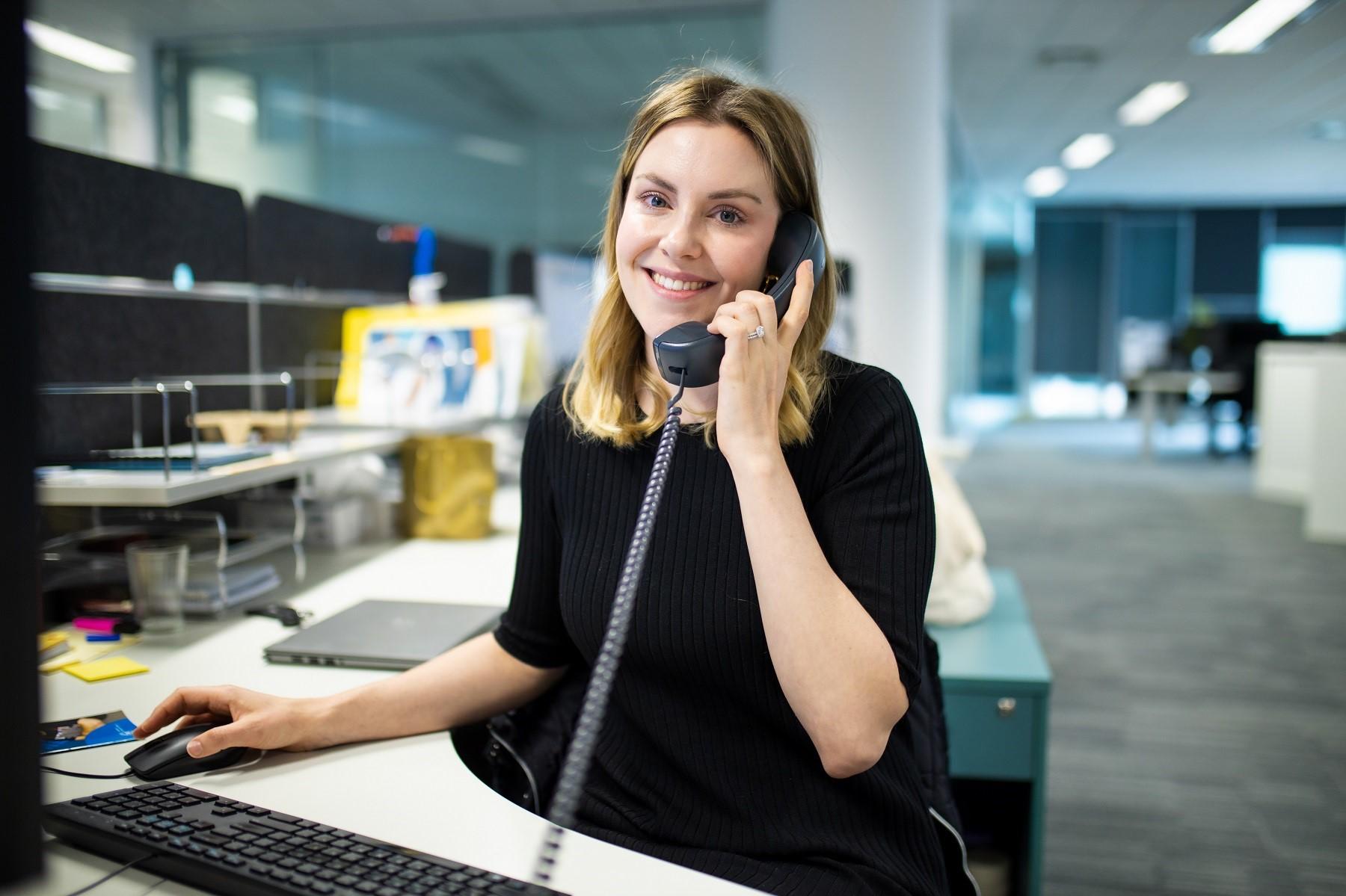 Photo of a woman smiling at the camera, on the phone sitting behind a workstation and computer.