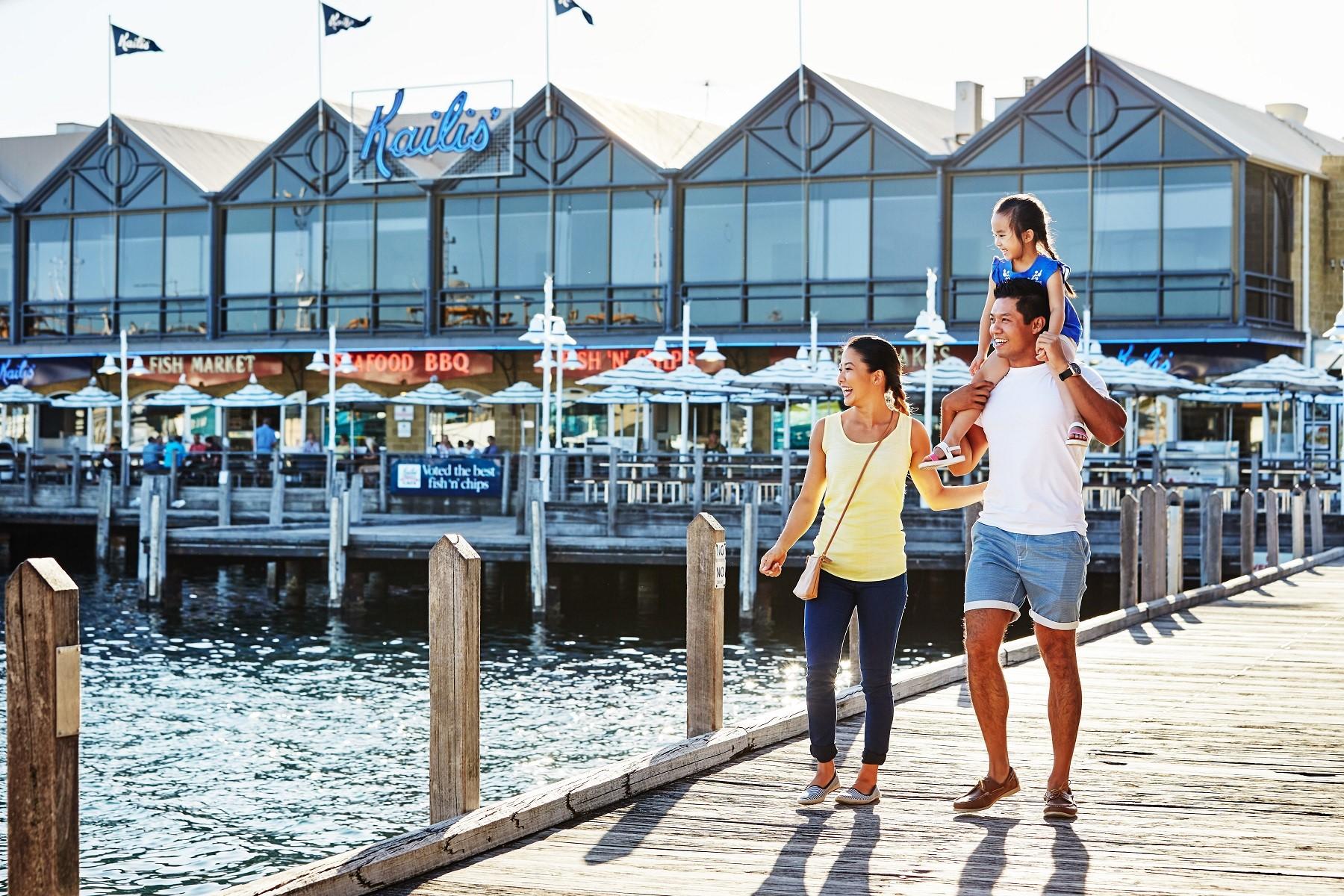 Photo of a man, woman and child walking along a jetty at Fremantle Boat Harbour with Kailis Fish Market in the background.