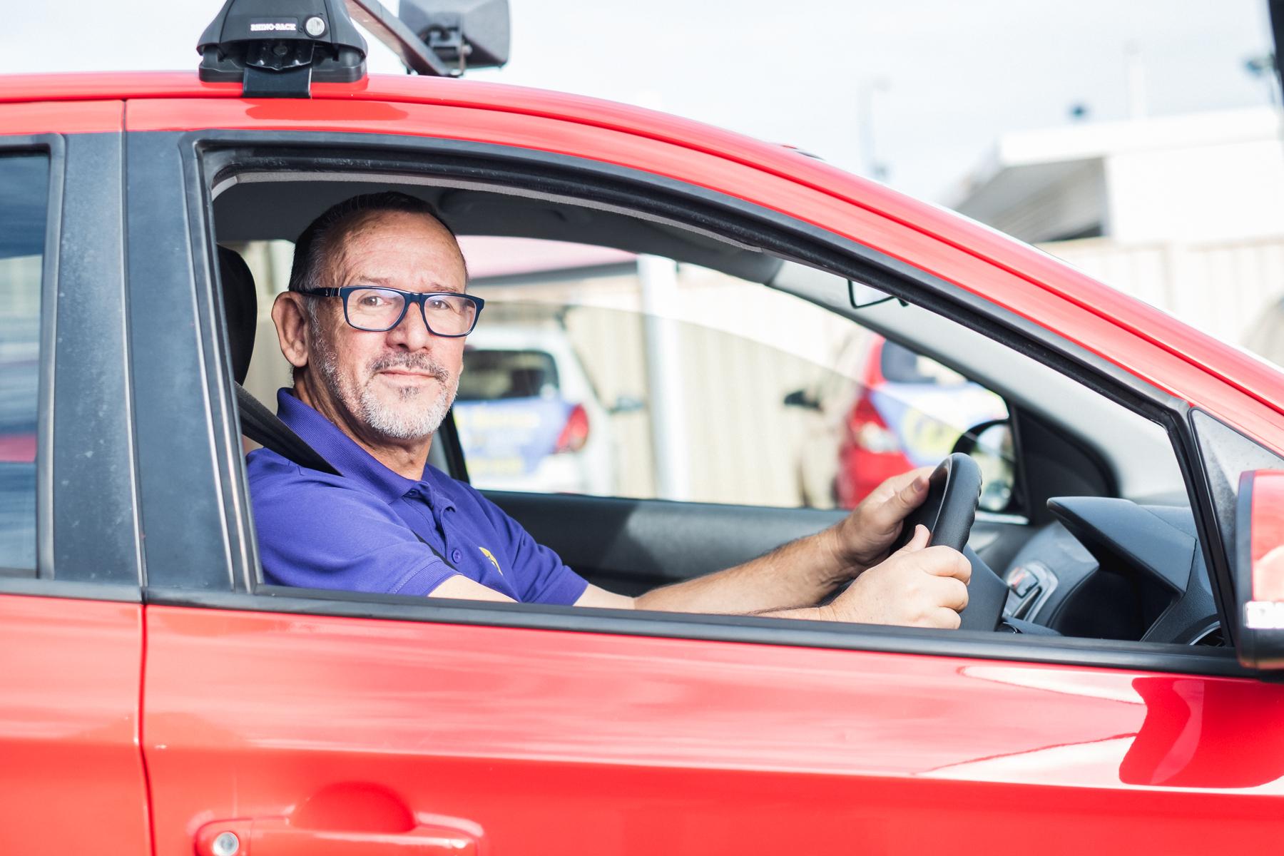 Image of a man sitting in a car, turned, looking out the car drivers window to the camera.
