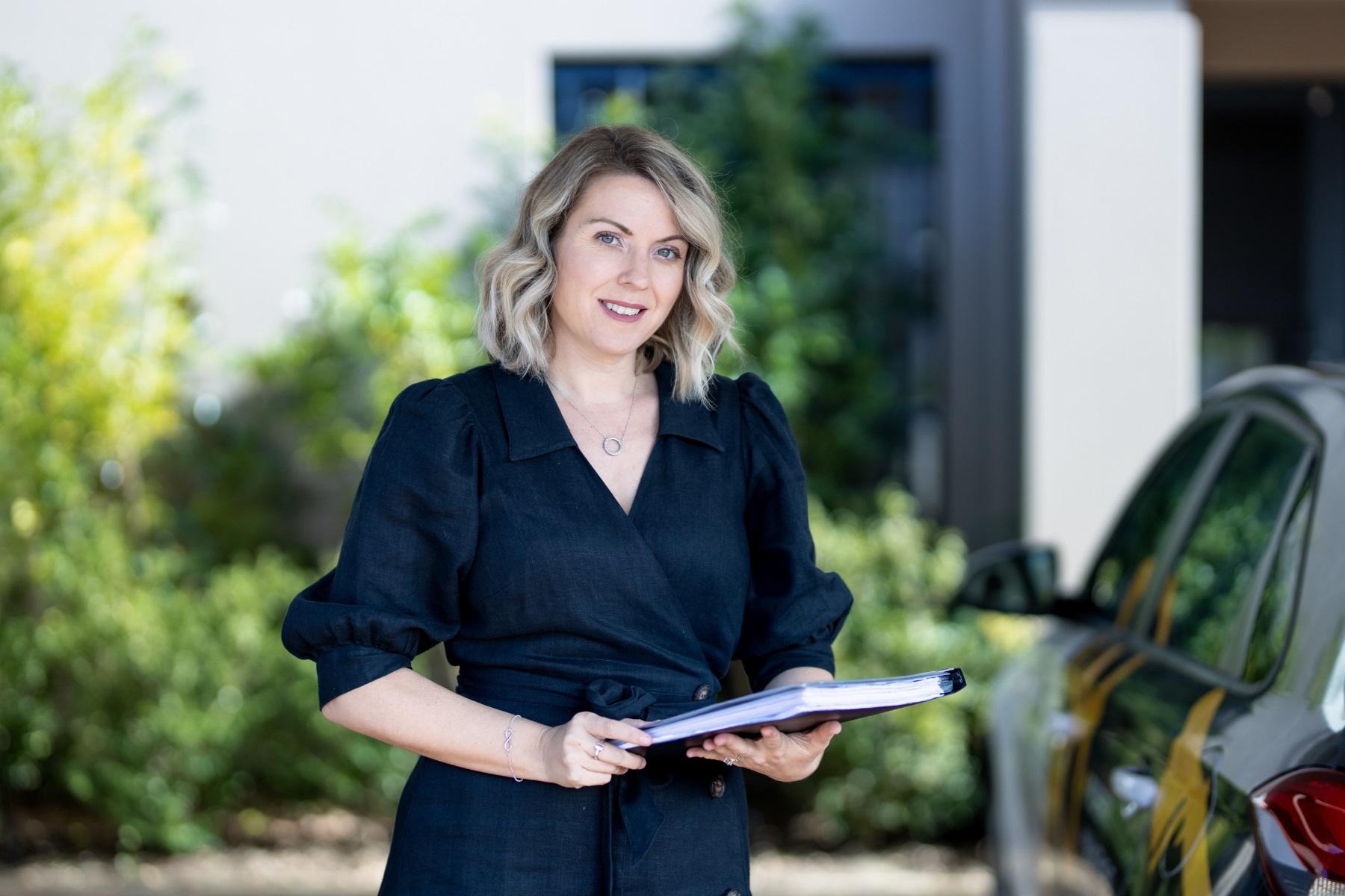 Woman standing by a car with a notebook in her hands.
