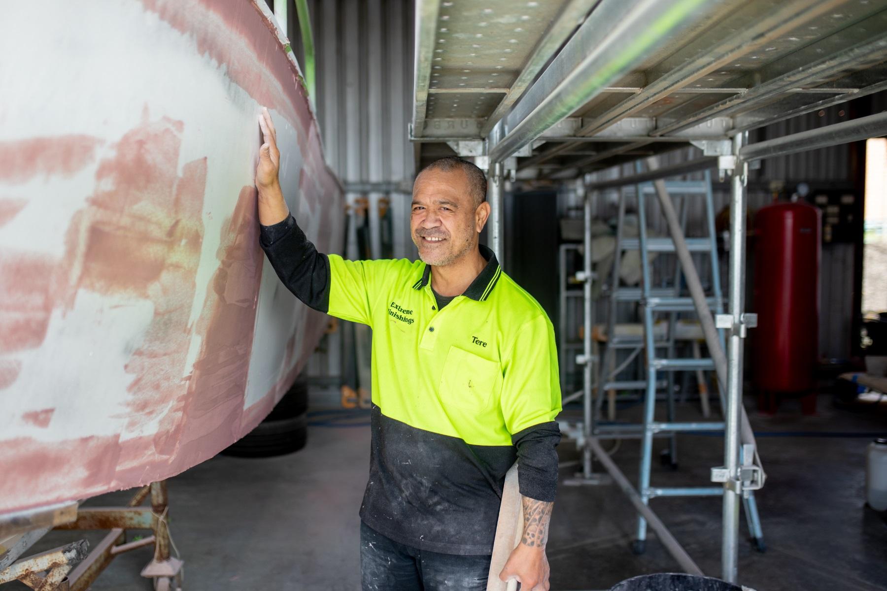 A man in a workshop standing next to the hull of a boat.