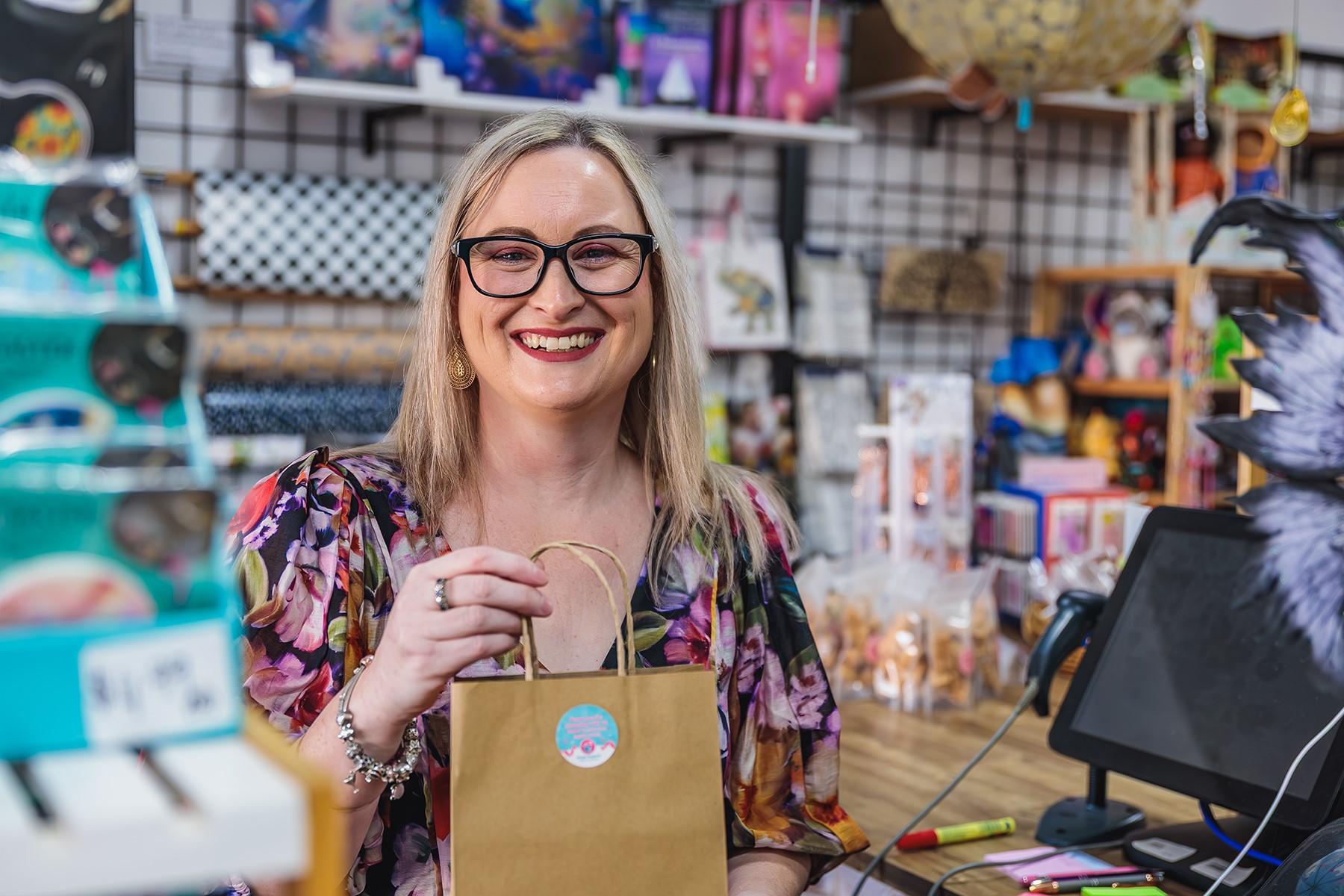 A woman standing at the counter of a retail store. She is holding a bag and smiling at the camera.