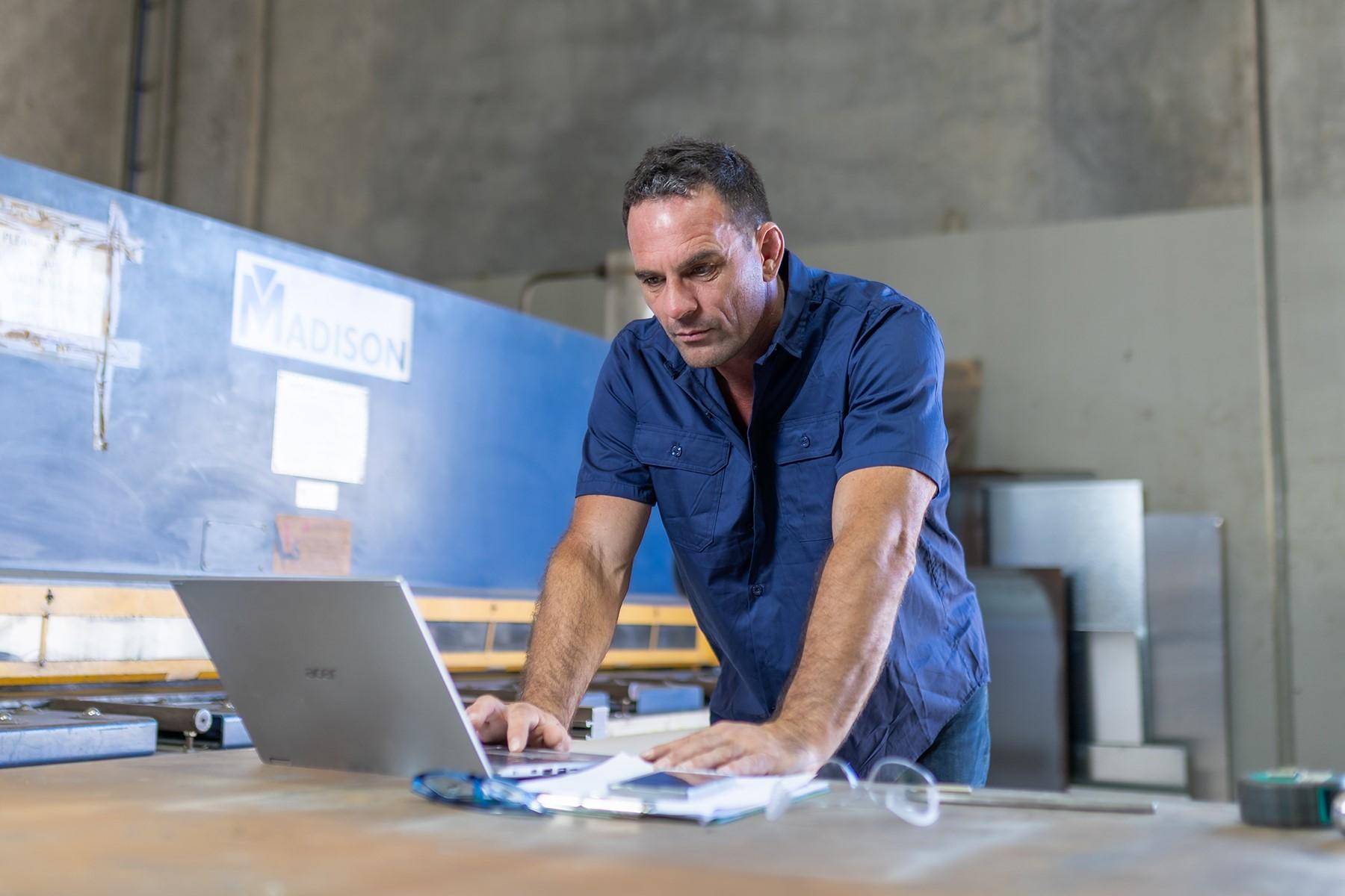 A man standing in an industrial workshop. He is standing at a workbench, reading from a laptop screen.