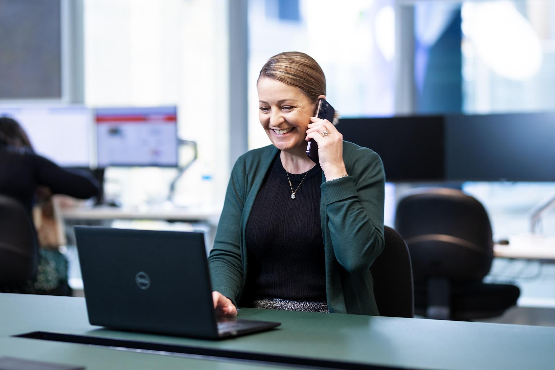 A photo of a woman sitting at a desk.She is using a laptop and talking on a smart phone.