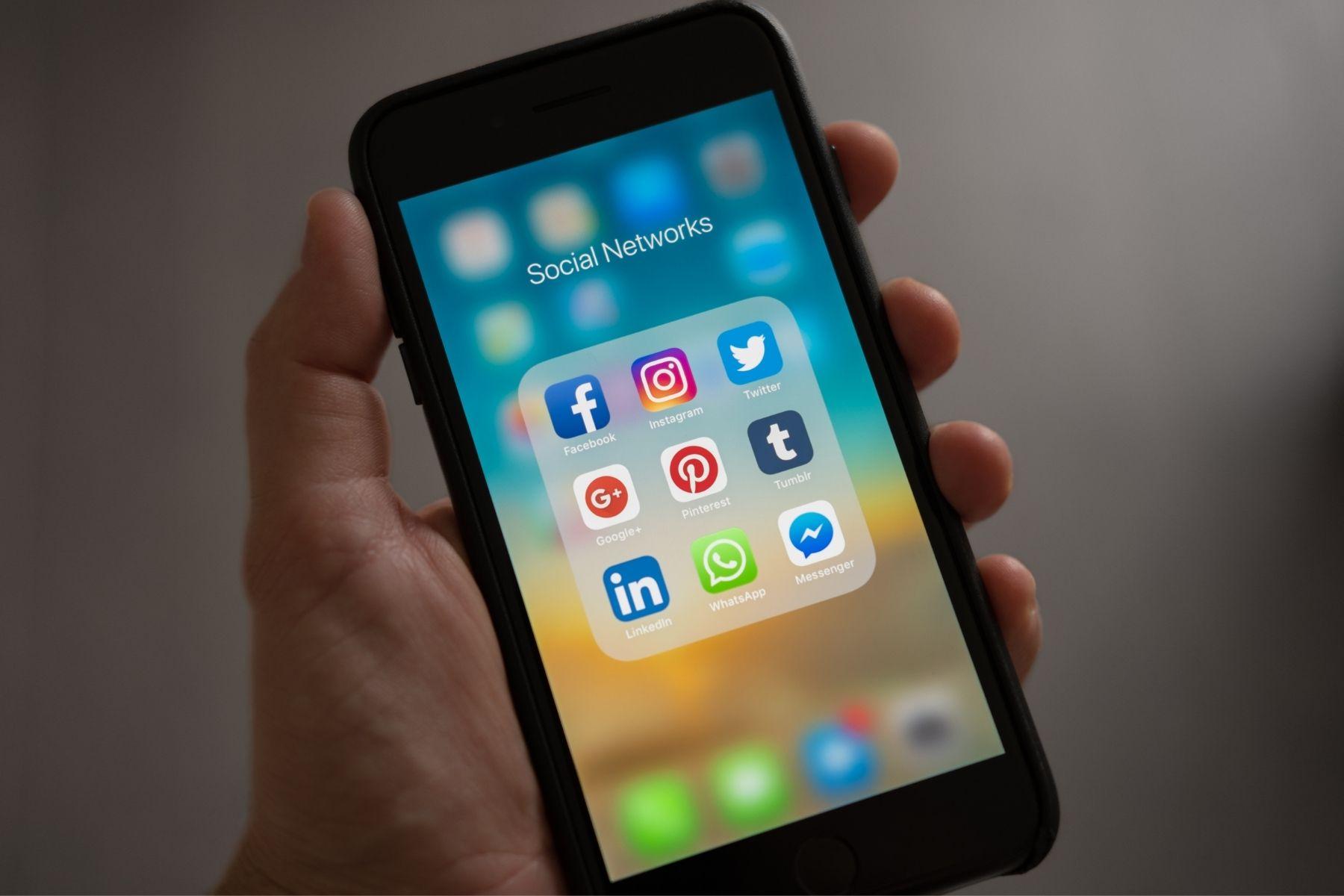 Photo of a hand holding a smart phone. The phone screen is displaying social media icons such as Facebook, Instagram, Twitter, and LinkedIn.