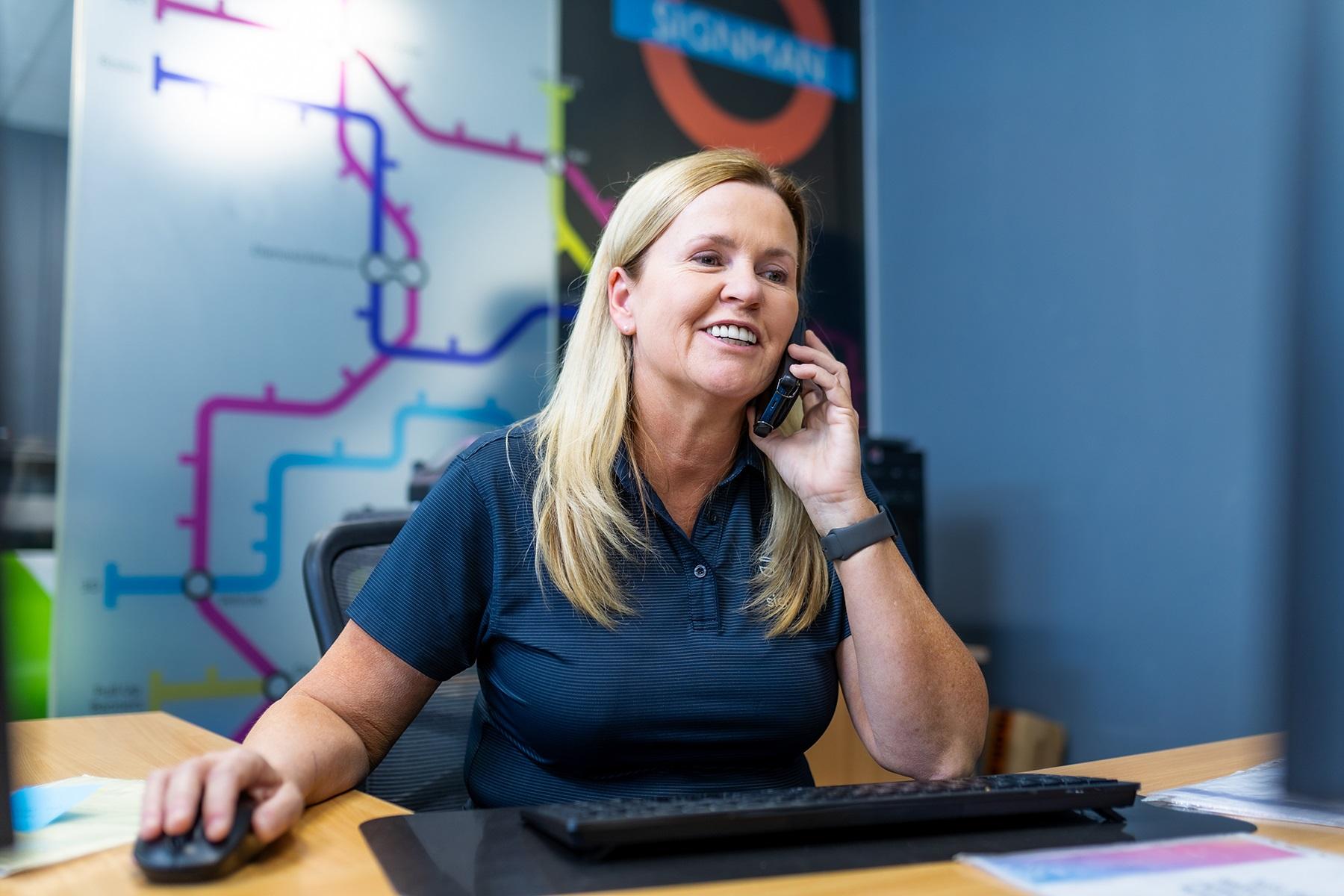 Photo of a female business owner. She is sitting at her desk and using her computer while she talks on the phone.
