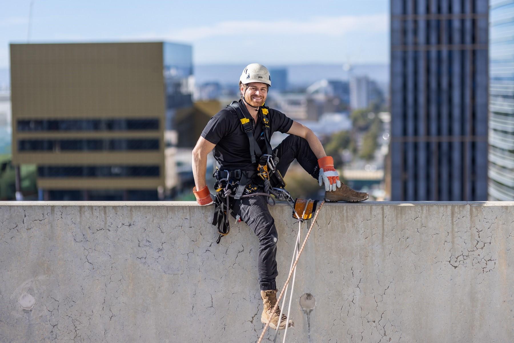 Photo of Alex Butchart, owner of High Access Window Cleaning. He is standing on top of a building and is wearing a hard hat and a harness.