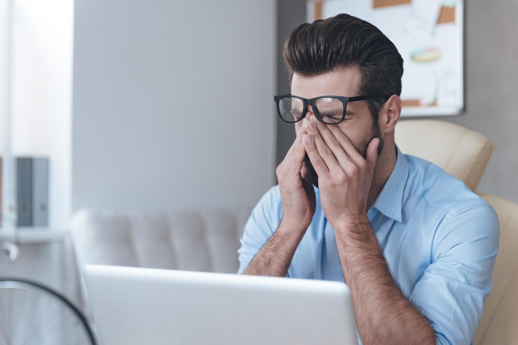 Photo of a man sitting in front of a laptop. He is rubbing his eyes.