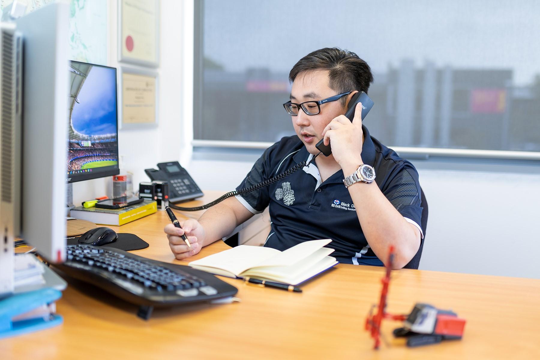 A man sitting in an office. He is speaking on the telephone.
