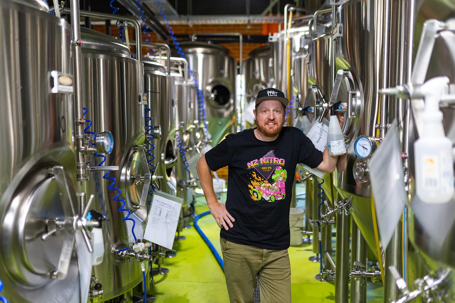 A man standing in a commercial brewery with distilling equipment in the background.