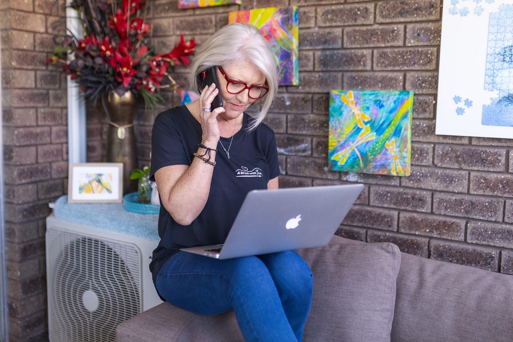 Photo of a woman sitting on the arm of a sofa. She is speaking on the phone while looking down at her laptop.