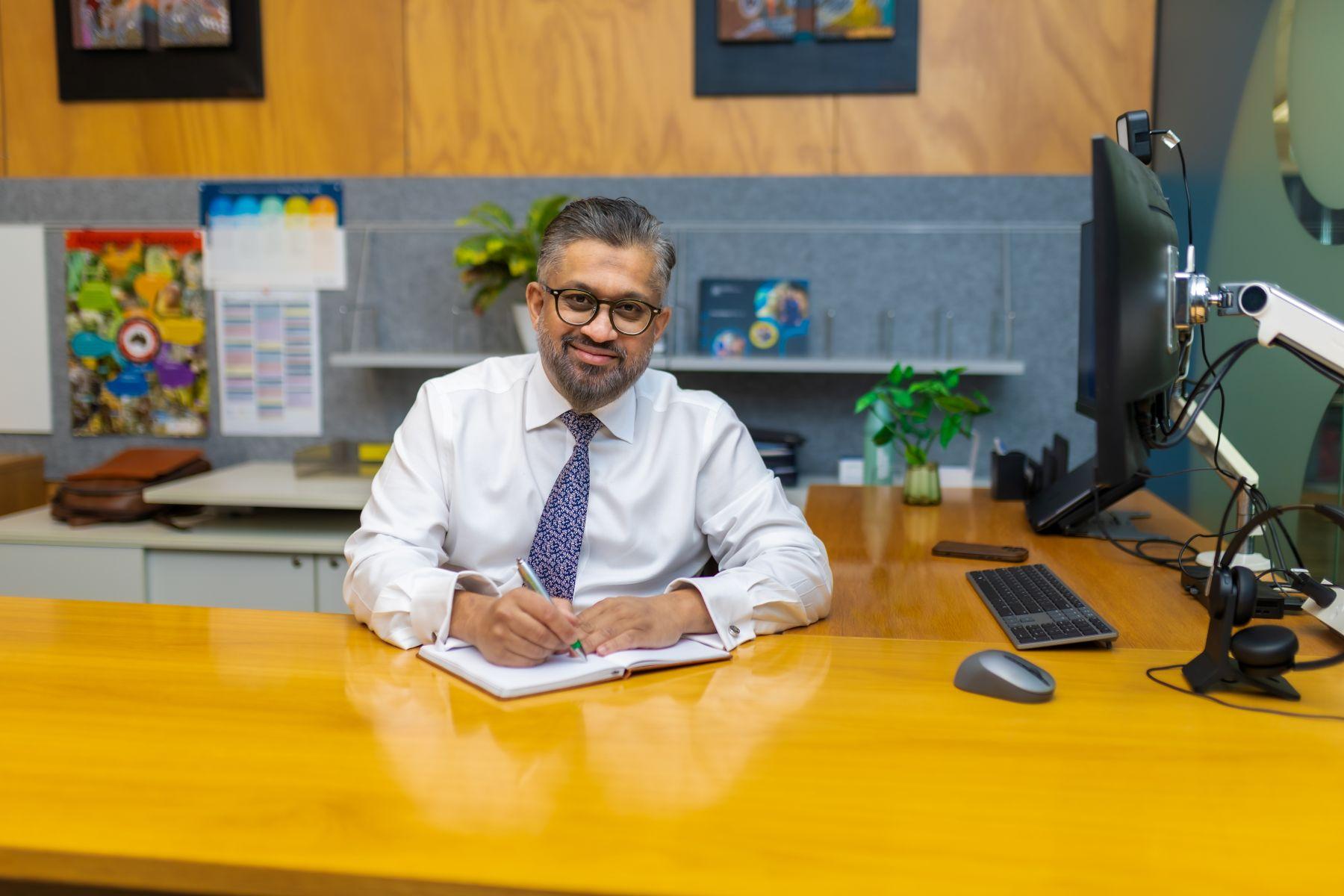 A man sits behind a desk, he is wearing a white shirt and tie and smiling at the viewer.
