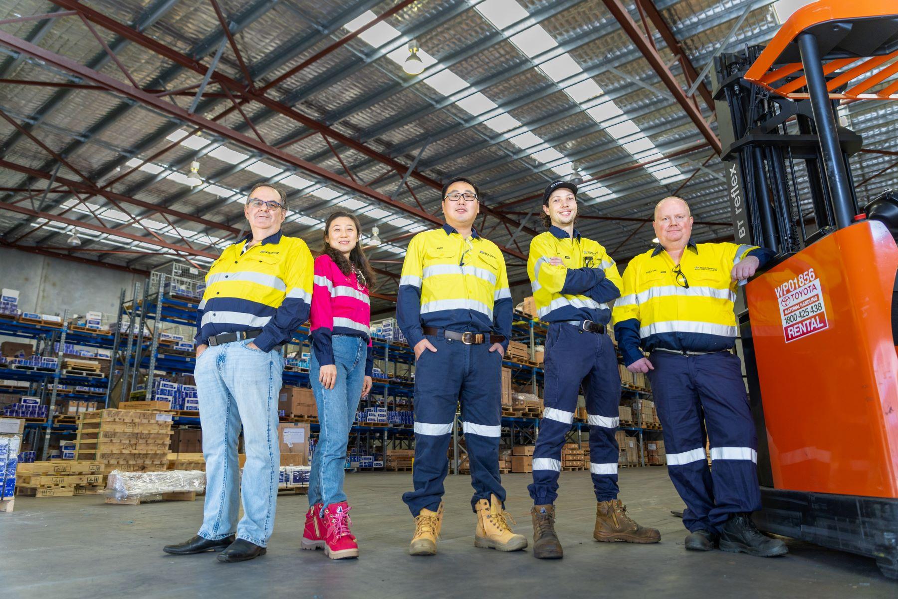 A group of five workers wearing high vis shirts are standing in a warehouse. To the right of a picture there is an orange pallet loader and there are shelves filled with boxes in the background.
