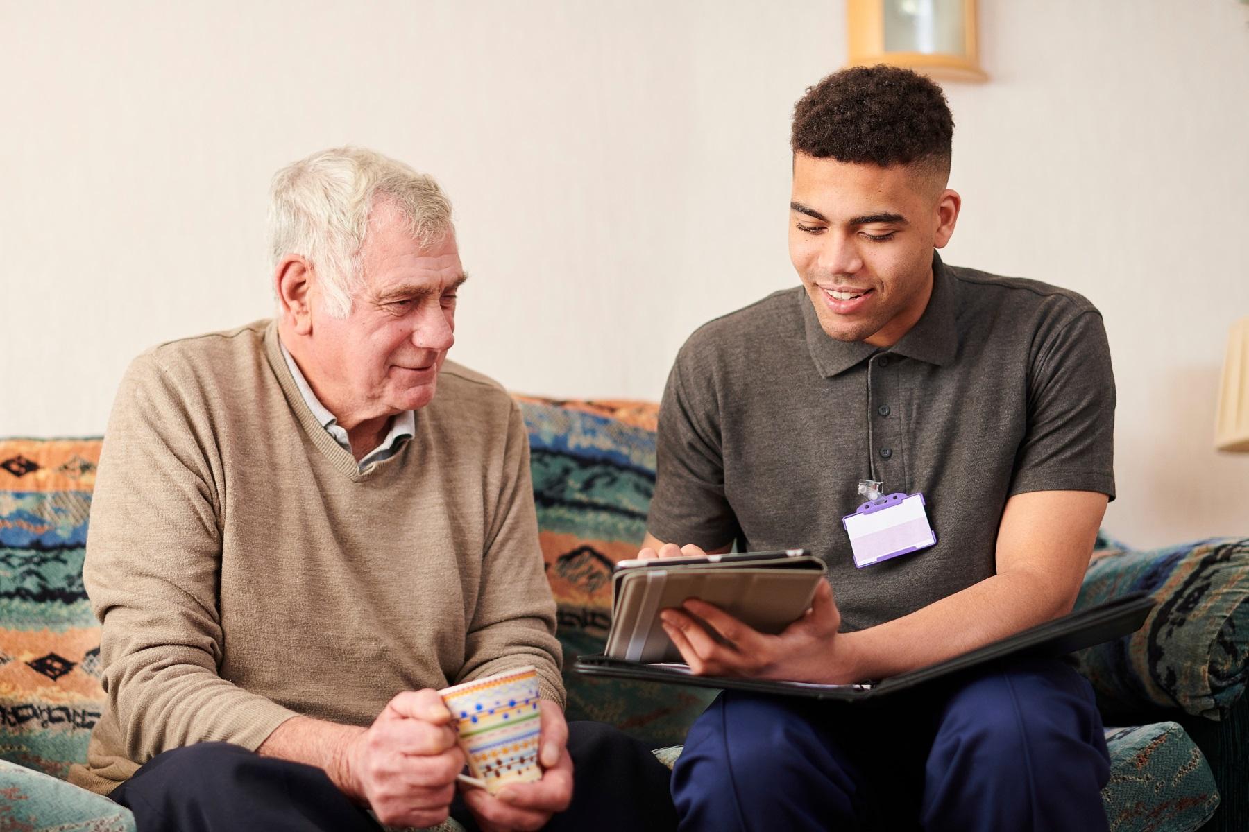 Photo of an elderly man being assisted by a male support worker. They are sitting together on a sofa and looking at a tablet device.