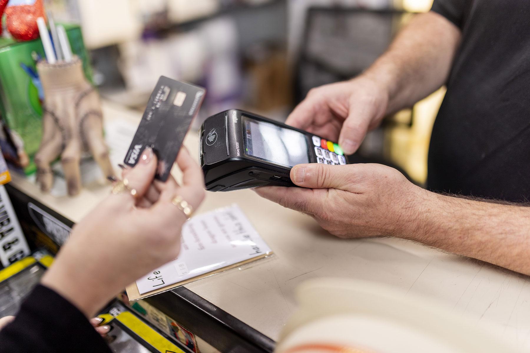 A shop owner holds out a card machine, and a customer is tapping their payment card.