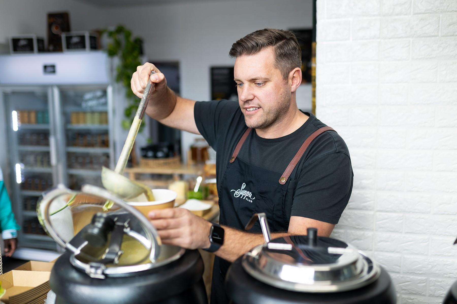 Photo of a male business owner ladling soup into a take away container.