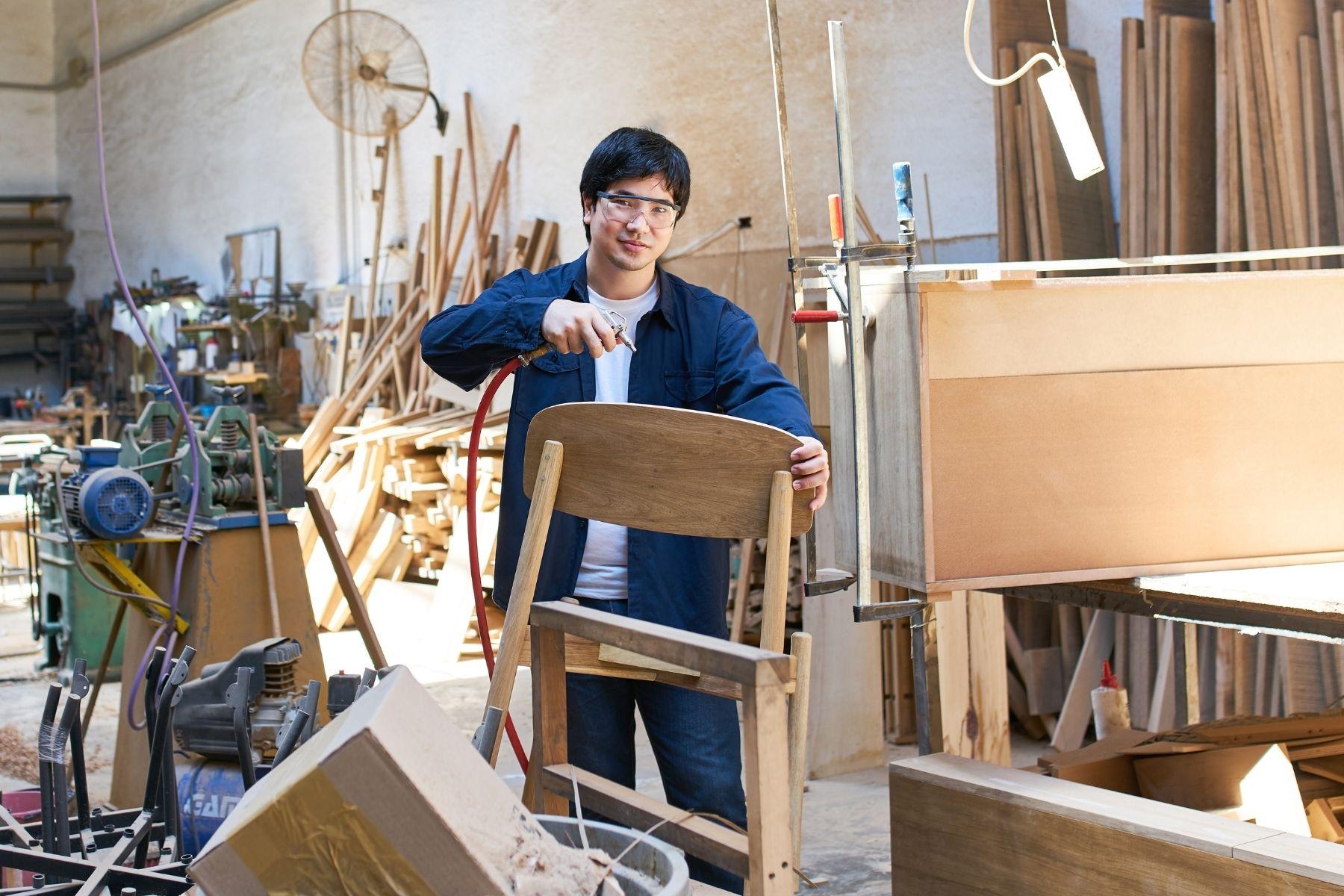 Image of a man in a furniture workshop spraying a chair, looking at the camera