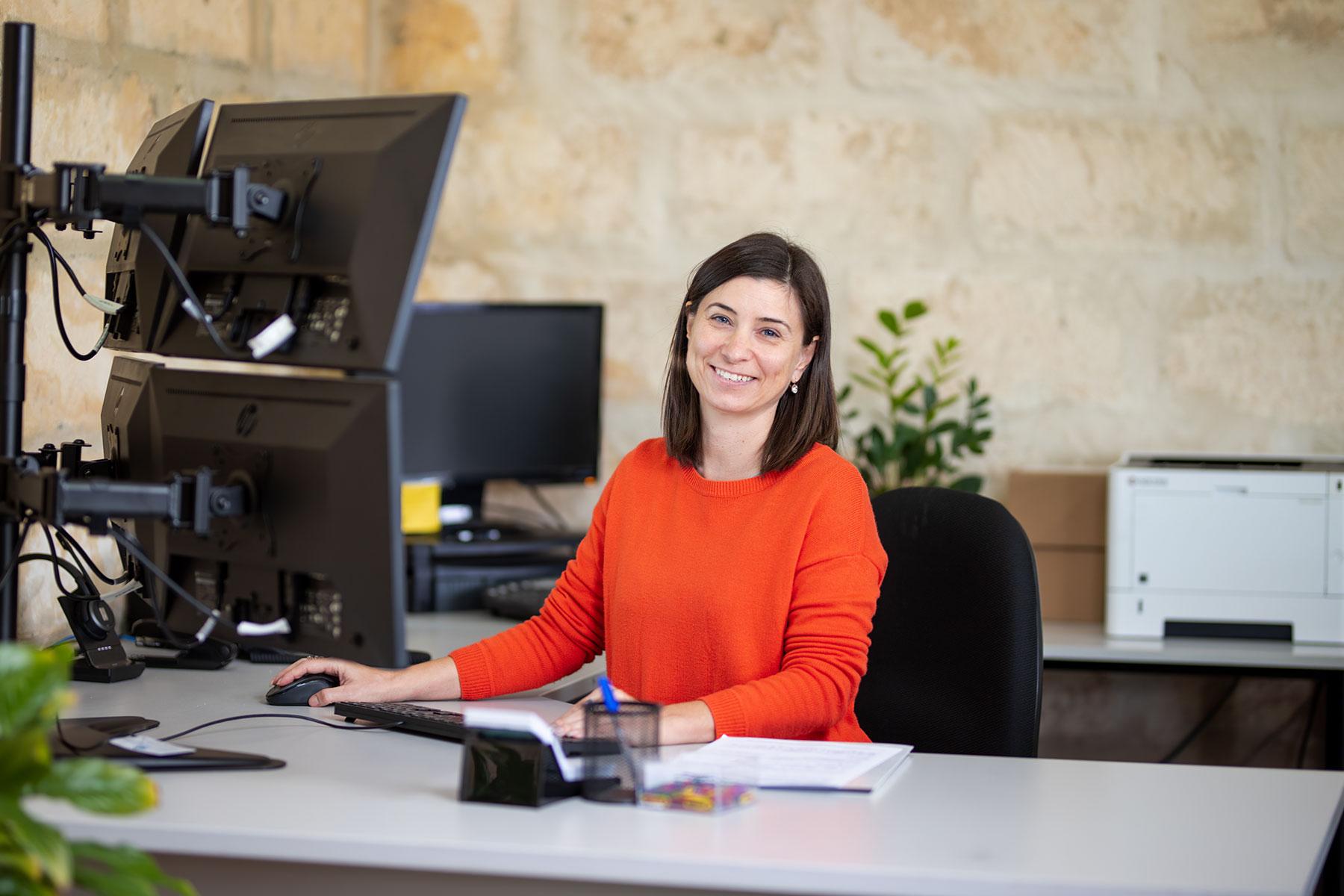 Image of a woman smiling at the camera sitting at a desk in front of computer screens