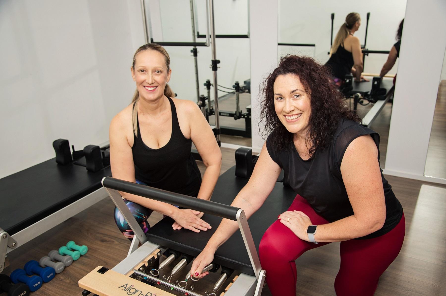 Image of two ladies adjusting the springs on a pilates  reformer bed, smiling at the camera.