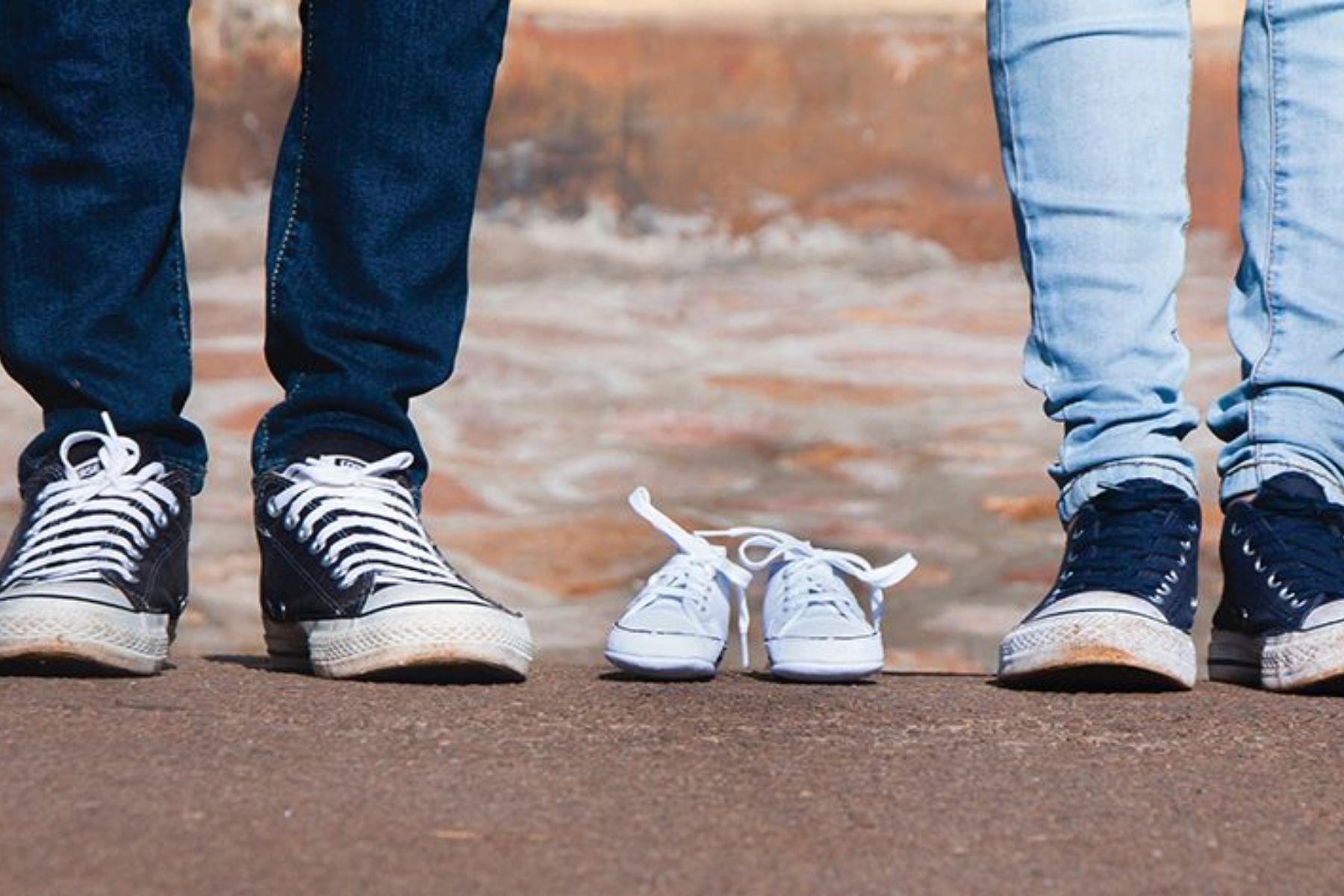 A close up photo of a man and women's feet and legs with an empty paid of baby shoes between them.