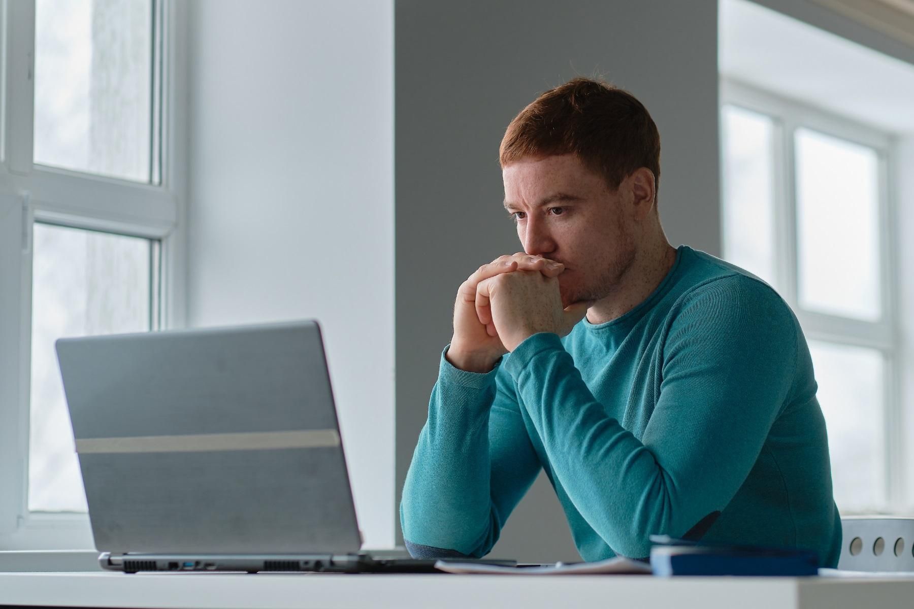Photo of a man sitting in front of a laptop. He has his hands to his face and looks concerned.