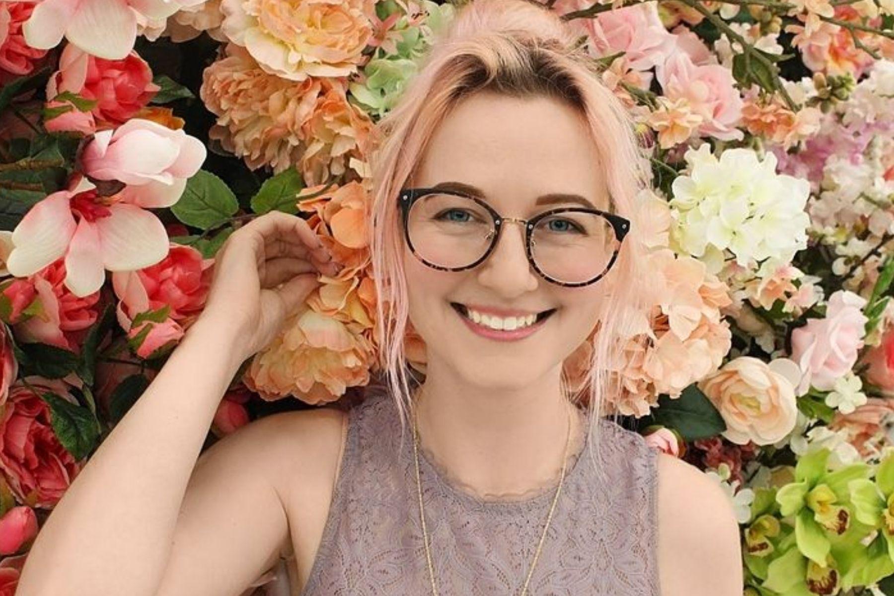 Photo of a young woman standing in front of a wall of flowers.
