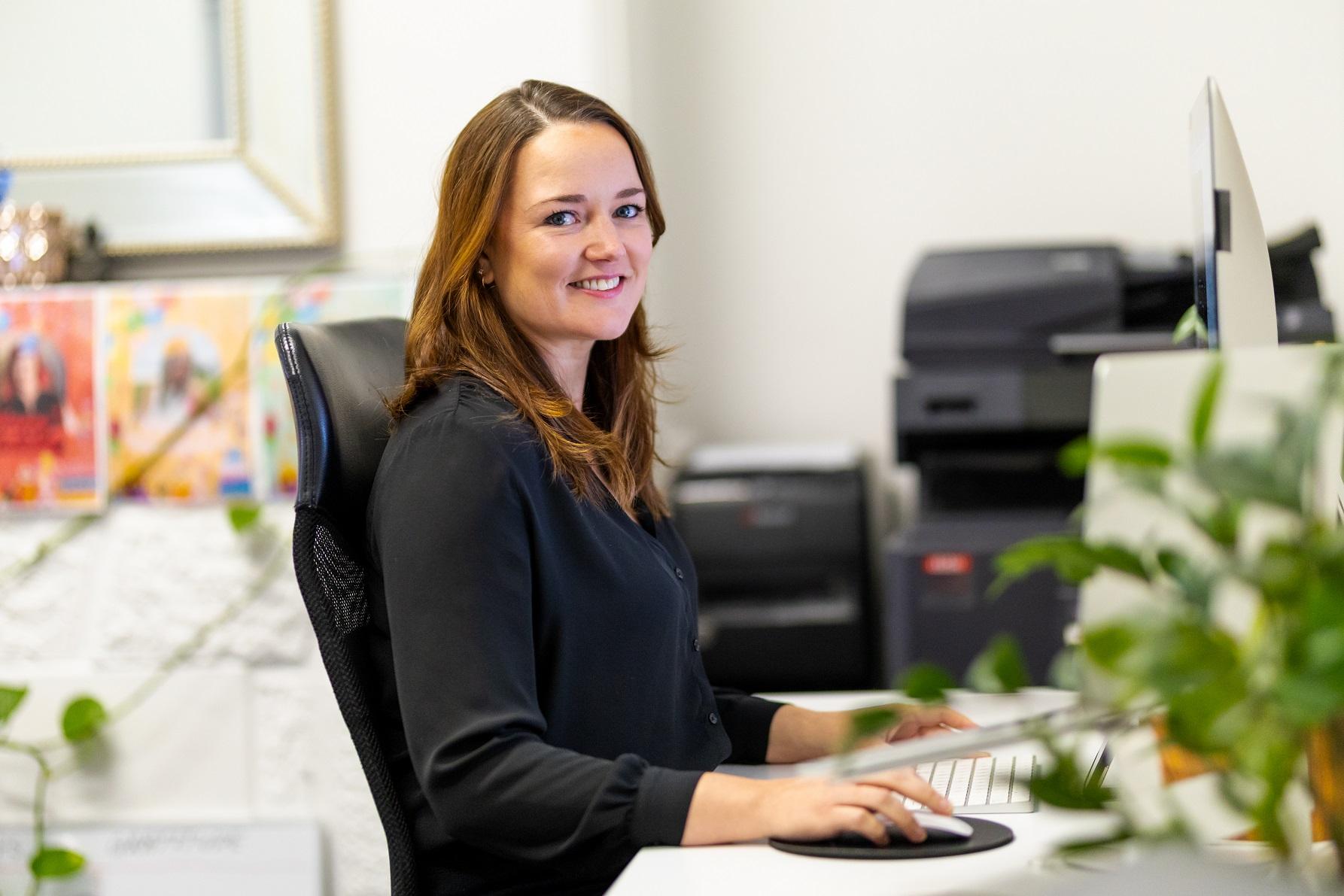 Picture shows lady sitting at a desk with her keyboard in from, turning to face the camera and smiling